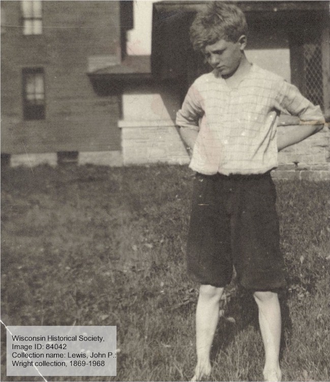 PHotograph of boy in striped, long-sleeved shirt and shorts in summer, with buildings behind him.