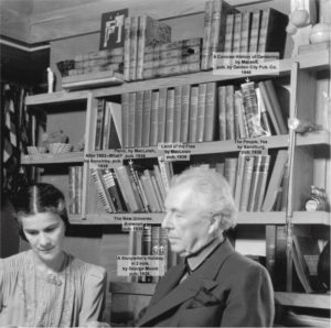 Photograph of Frank and Olgivanna Lloyd Wright in front of a bookshelf at Taliesin. Some of the books are named.