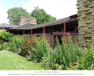 Color photograph taken of outside garden. Showing grass, foliage, and red metal trellises that Wright designed.