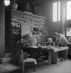 Photograph of Frank Lloyd Wright at his desk. Taken in 1957. Eugene Masselink is standing with him.