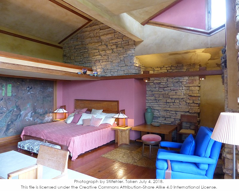 A photograph looking north in Taliesin's Guest Bedroom taken while on a tour. Includes the bed, several seats, and lamps. Has masonry in view. Photograph by Stilfehler.