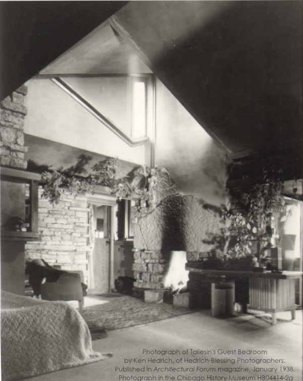 Photograph of Taliesin's Guest Bedroom taken by Ken Hedrich. Taken in 1937 Has a bed, chairs, furnishings, and a wooden door. Fire in the fireplace.